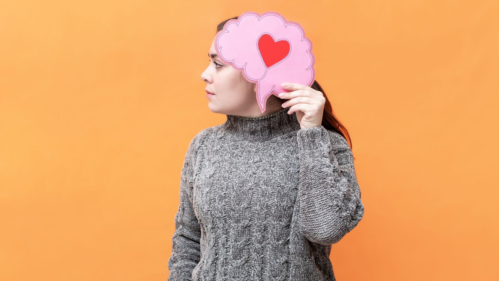 Image representing mental health care, self care, and self love with woman holding piece of paper with a heart on it over face